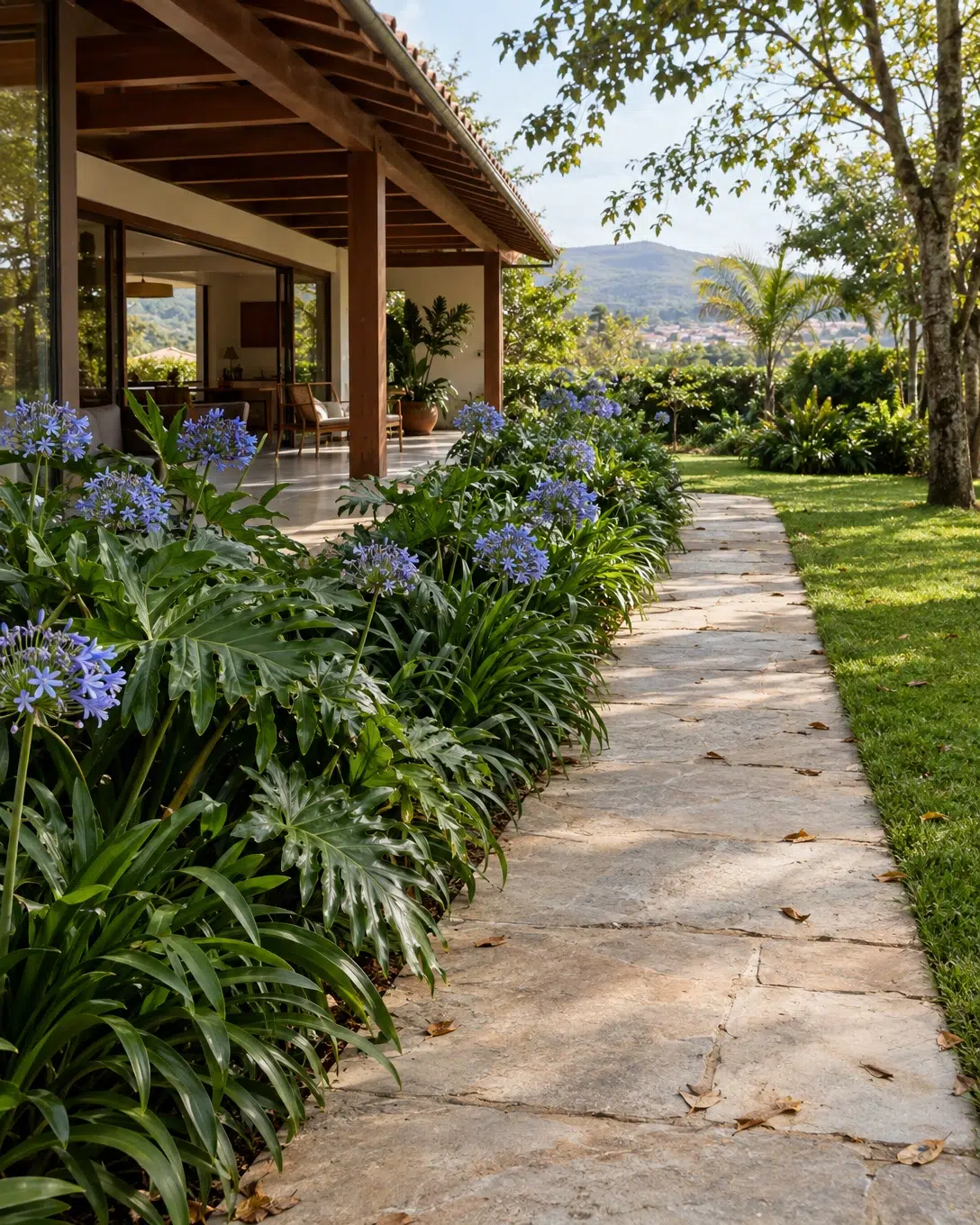 Decoração de chácaras e sítios: como deixar o espaço charmoso 1 Varanda de casa de campo com piso de pedra clara, ladeada por canteiro largo com agapantos e gramado ensolarado.