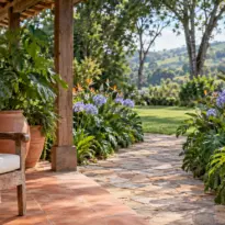 Varanda rústica com poltrona de madeira e vasos de barro, aberta para jardim florido com caminho de pedra e morros verdes.