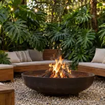 Espaço de estar em quintal com fogo de chão central sobre pedriscos, bancos de madeira e folhagens tropicais ao fundo.